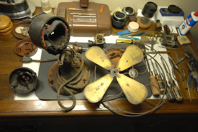 Color photograph of an antique fan disassembled on a workbench with motor, guard, blades and hand tools.